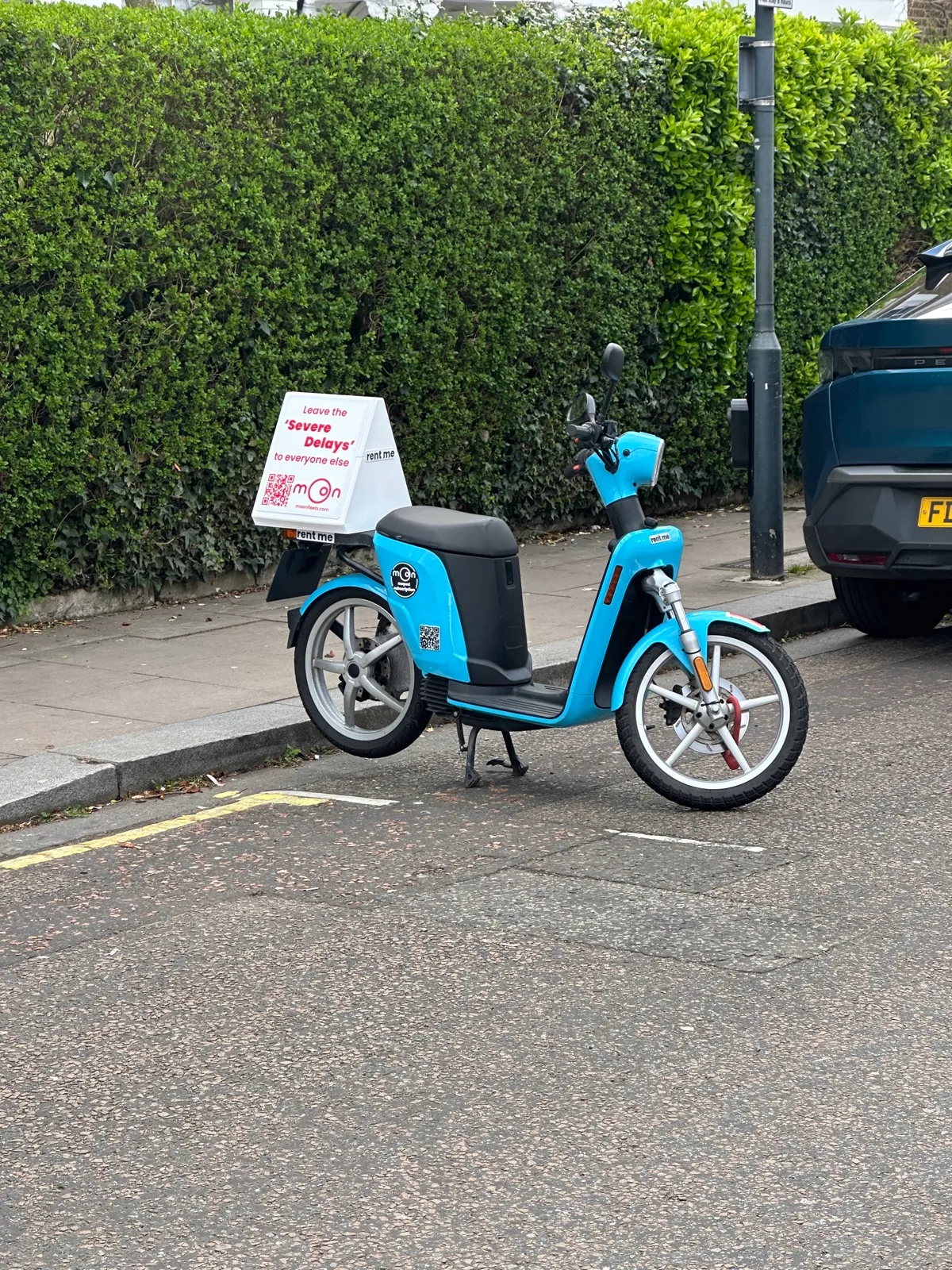 A sky-blue Moon Fleets Askoll electric moped parked kerbside in London, carrying an ad box that reads: Leave the ‘Severe Delays’ to everyone else